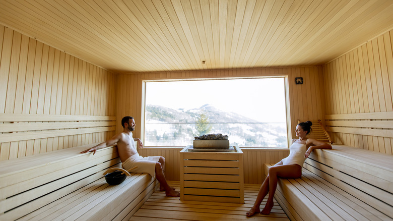 A couple in white robes sits in a wooden sauna overlooking a winter landscape in Finland, smiling