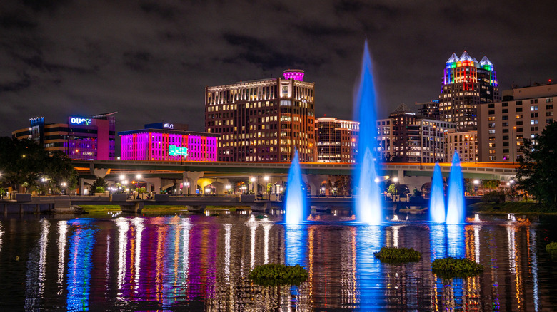 Illuminated fountains glowing blue at night with downtown Orlando skyscrapers and buildings lit in vibrant colors, reflected in the water below.