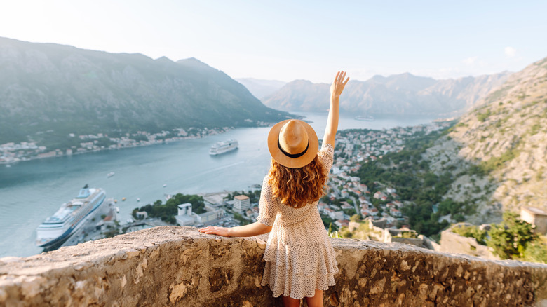 Back view of a young female tourist wearing a polka dot dress and a straw hat during a summer European trip, overlooking a waterway with cruise ships