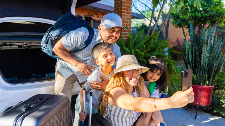 A family taking a selfie in front of an open car trunk, with luggage around them