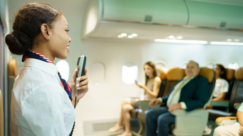 A flight attendant speaks into the PA on an airplane with seated customers in the background.
