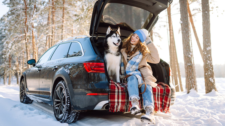 Woman and a dog sitting in the back of a car, with the trunk open, surrounded by snowy scenery