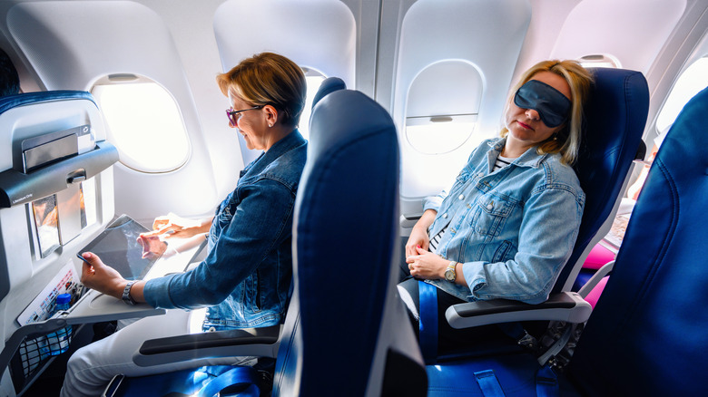 Two women sit on an airplane in separate rows, the one in front on an iPad, the one behind sleeping in an eye mask.