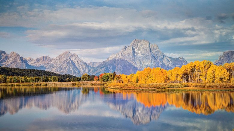 A view across a glassy lake in Yellowstone shows fall foliage and mountains in the distance.