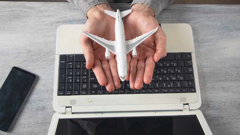 Hands holding a toy airplane above a laptop computer on a gray desk next to a black phone