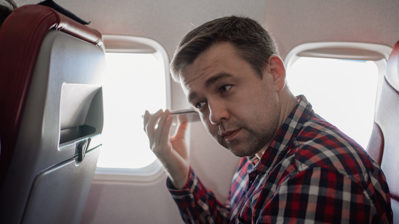 A man in a plaid shirt listening an audio message on a phone, while sitting on a plane.