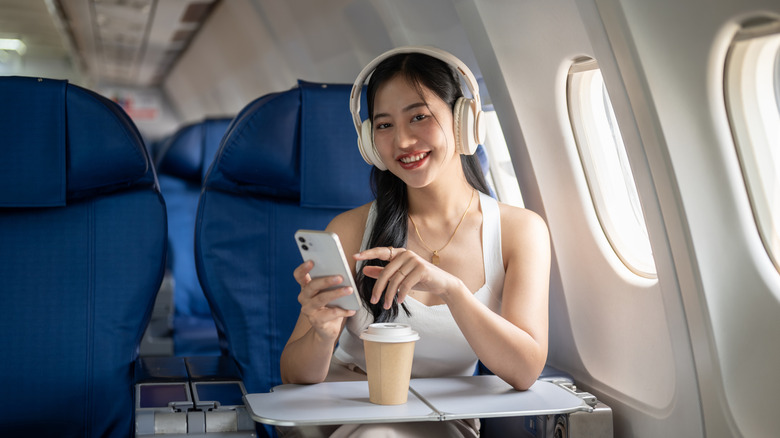 A smiling woman using her smartphone and wearing headphones during a flight.