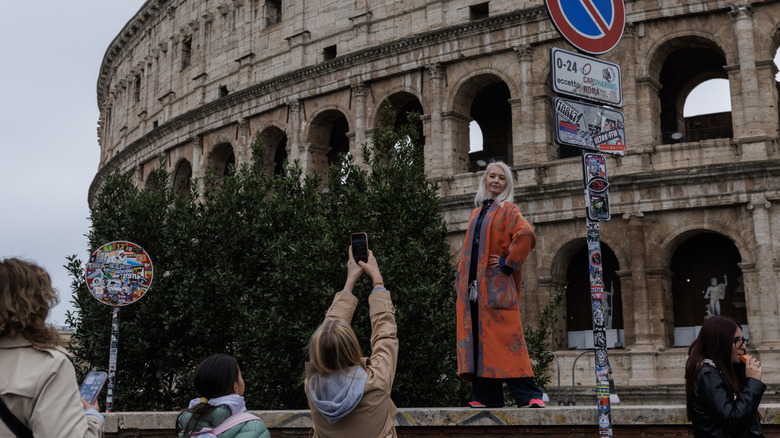 A woman poses for a photo while standing on ledge in front of the Colosseum.