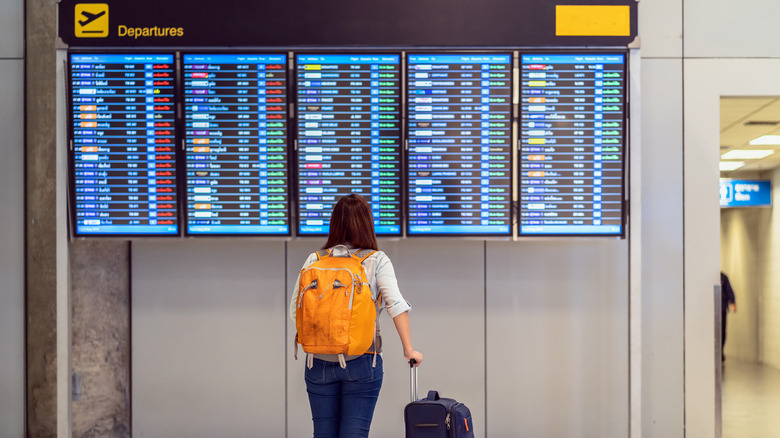 A woman stands with her luggage, facing the flight information board at an airport.