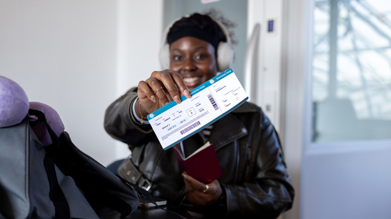 A close up of a female solo traveller who is holding up her airline ticket and smiling as she looks into the camera.