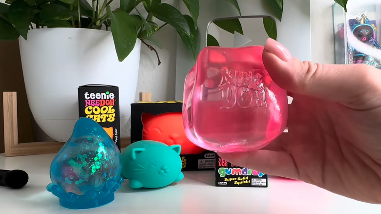 A hand holding a NeeDoh squishy toy, with other similar toys in the background on a flat surface