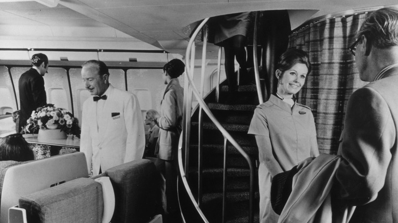 An air hostess greets passengers at the base of a spiral staircase on an airplane circa the 1970s.