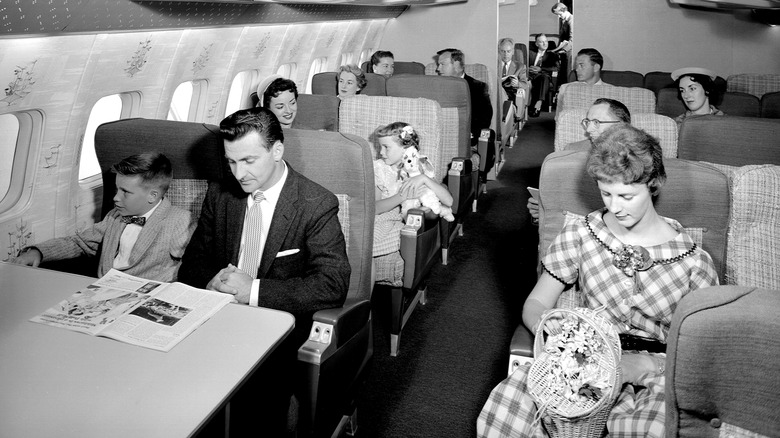 Passengers pictured on an airplane, including a woman wearing a dress, and a man wearing a jacket and tie.