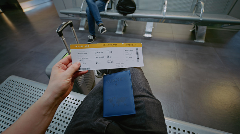 First-Person View of Traveler with Suitcase, Passport and Boarding Pass Sitting in Departure Lounge of Airline Hub