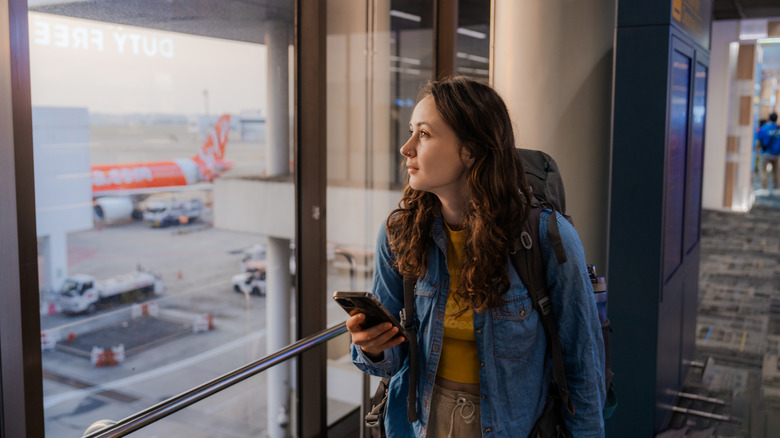 Woman wearing backpack gazes out at the airport tarmac with a cellphone in her hand.