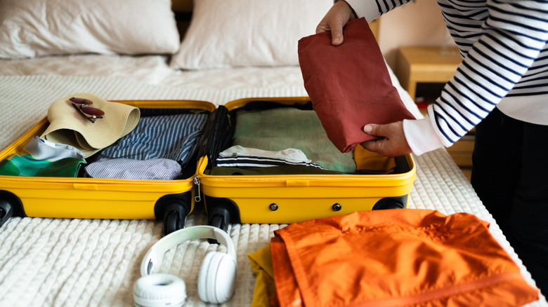 A person placing objects in a suitcase sitting on a bed