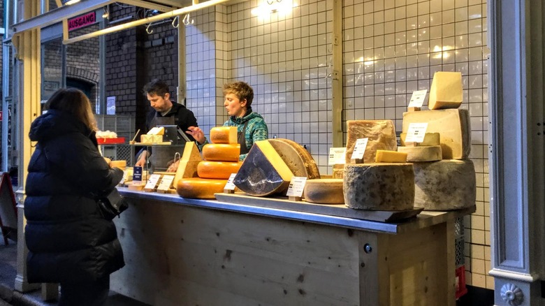 A woman at a checkout counter displaying large wheels of cheese