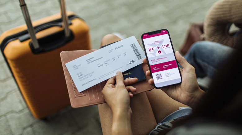 An airline passenger sits, holding their mobile boarding pass next to their printed boarding pass with luggage in the background.