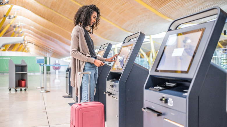 A woman with a pink suitcase checks in at an airport self-serve kiosk.