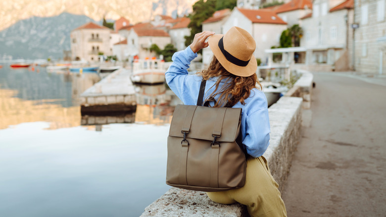 A woman wearing a hat and a backpack sitting next to the water in an old European city
