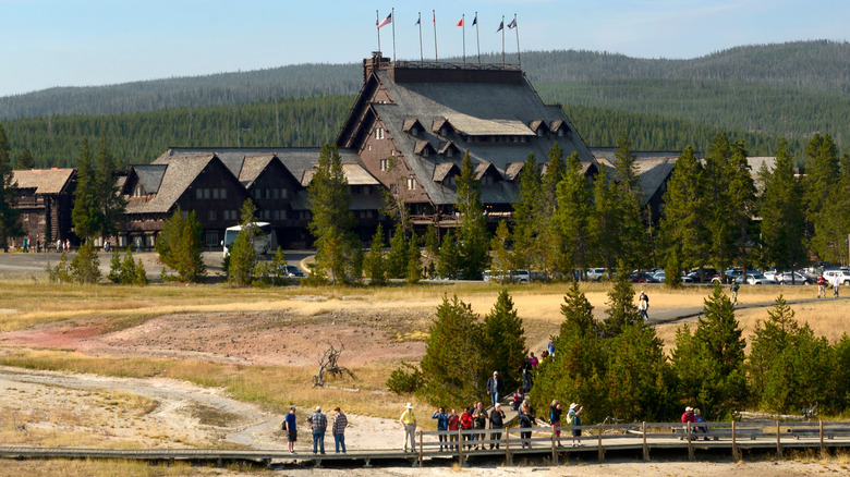 A large wooden lodge in front of evergreen hills, with tourists on a walking path