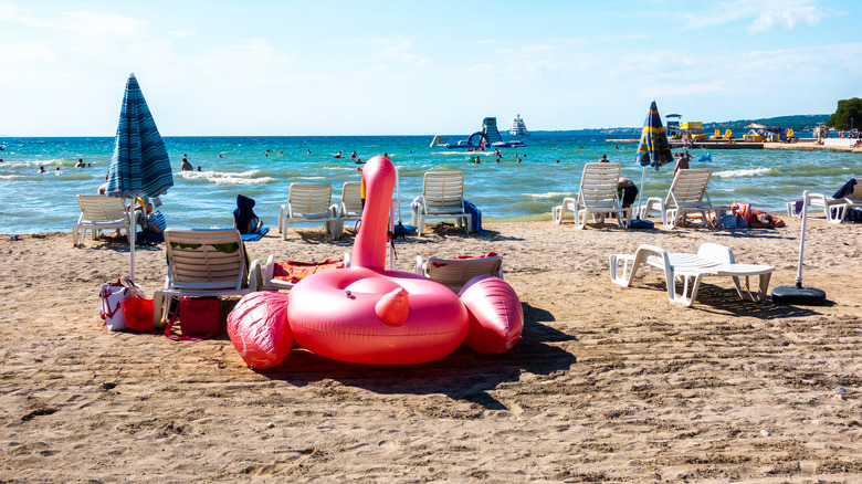 an inflatable flamingo sits on the sand among lounge chairs ocean in background